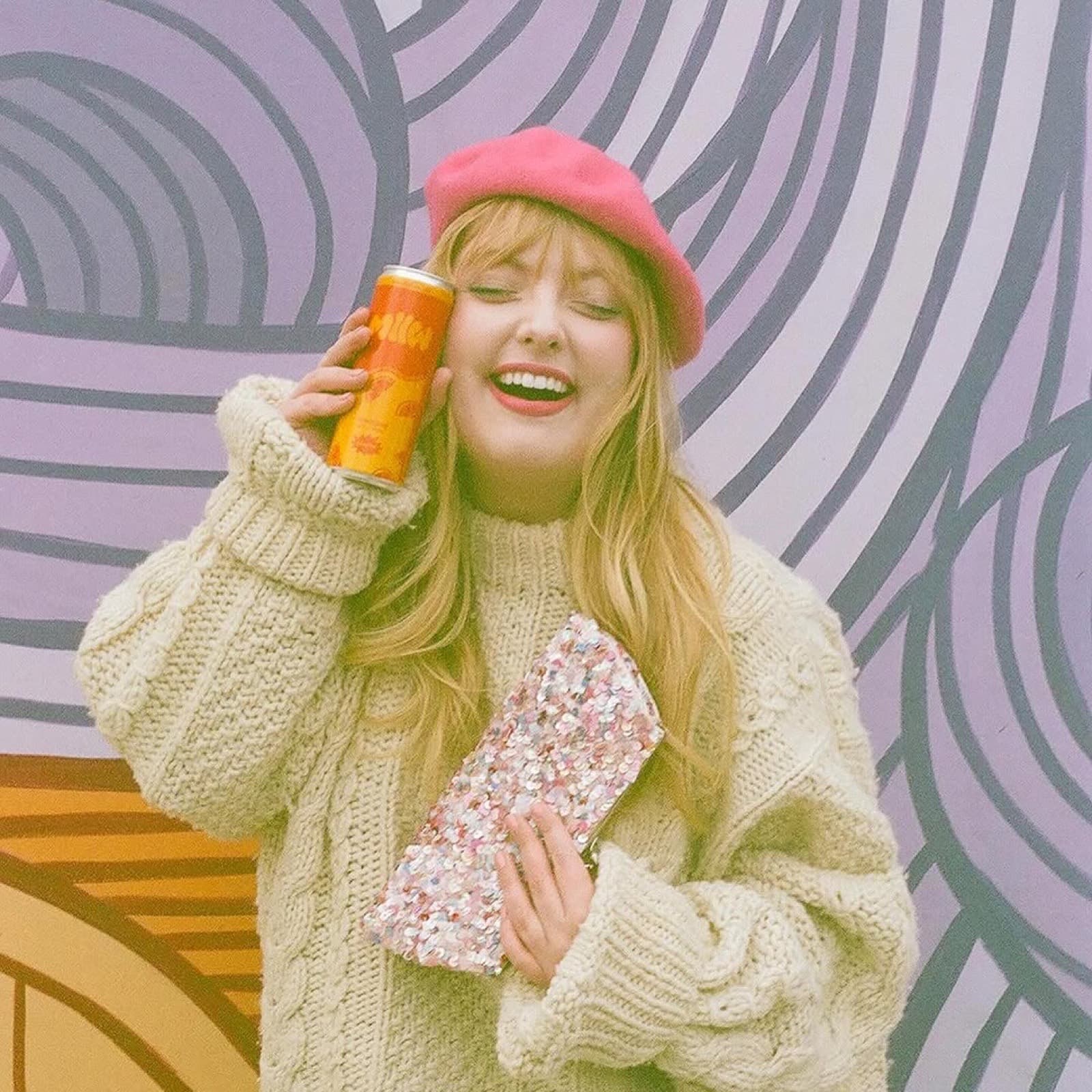 Woman in pink beret holding Tangerine MLLW can against swirly mural